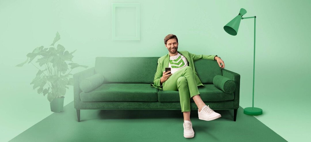 elearnio – Mood Bild Kunde smava Das Bild zeigt den bekannten "smava hero" wie er auf einer grünen Couch sitzt und in die Kamera lächelt. Das Bild ist in Grüntönen gehalten.