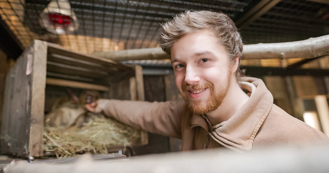 Das Bild zeigt einen jungen Mitarbeiter vom Erlebnis-Zoo Hannover mit hellen Haaren, Bart und brauner Kleidung vor einem Gehege.
