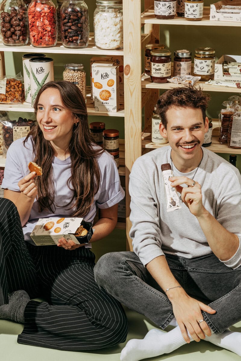 Auf dem Bild sieht man zwei junge Menschen vor einem Vorratsregal auf dem Boden sitzen. Sie essen Snacks der Marke "KoRo" - einem elearnio Kunden.
