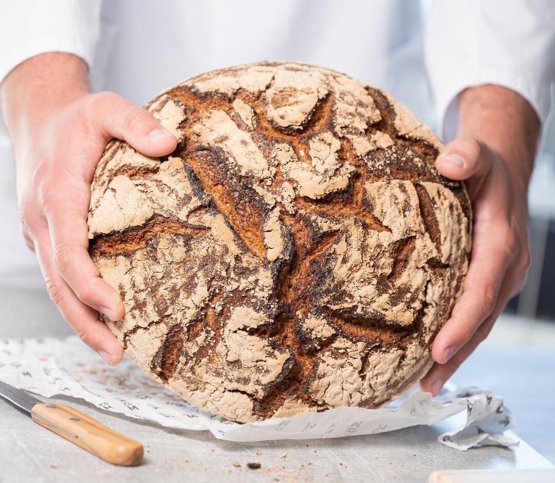 Nahaufnehme eines großen, runden Brotes – gehalten von zwei Händen einer Person, die nicht weiter sichtbar ist.