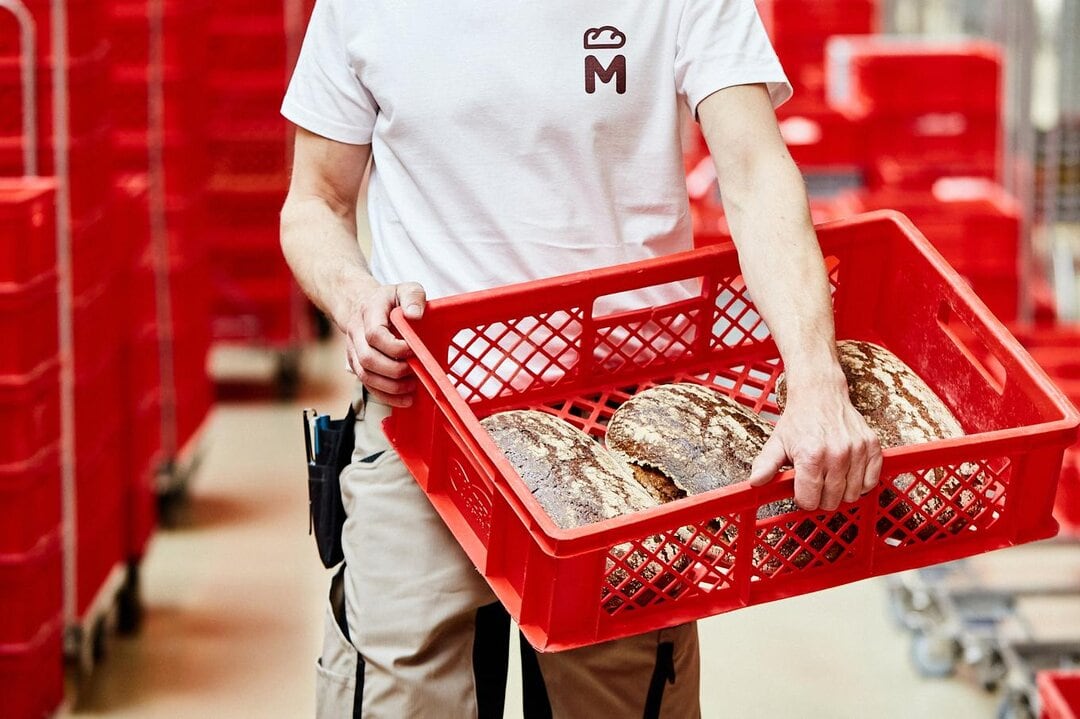 Person trägt in einer Produktionsstätte einen roten Korb mit Broten drin. Der Kopf und die Beine der Person sind vom Bild abgeschnitten.